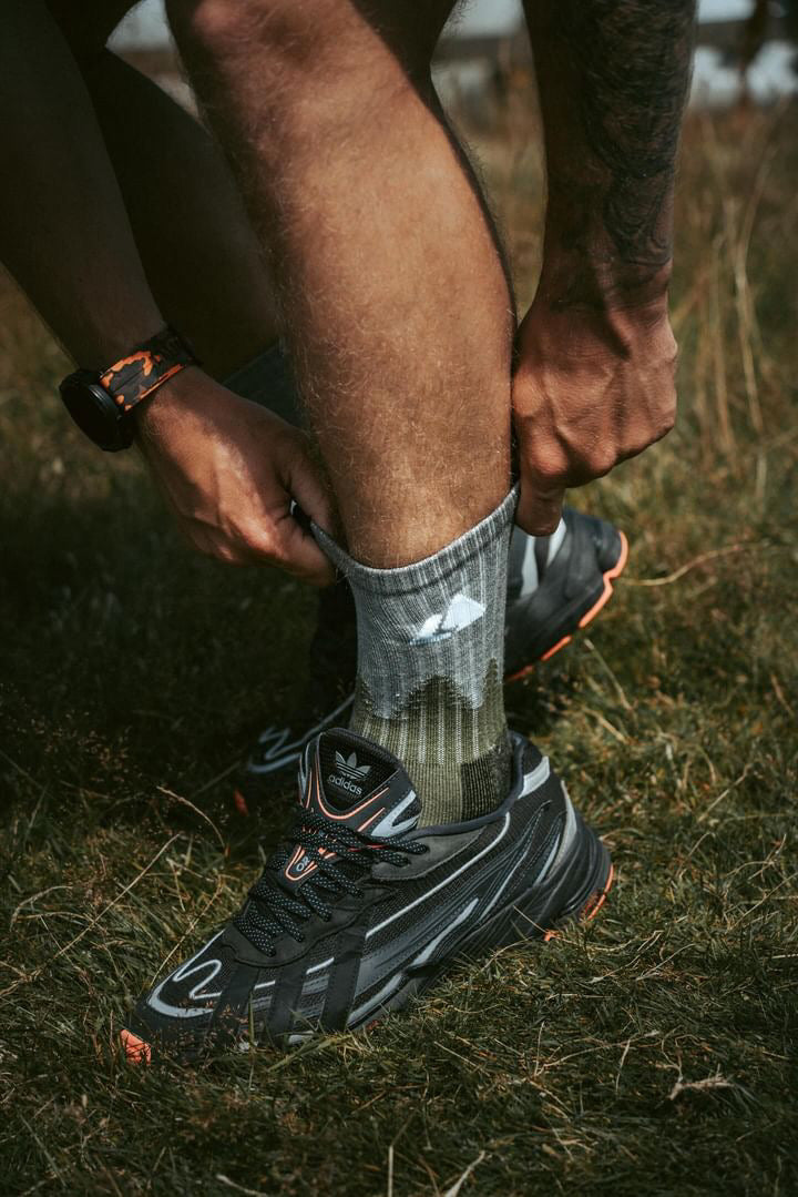 A pair of Merino wool hiking socks being worn on a person's legs, with one sock displaying a design or logo, in an outdoor setting with grass and a watch on the wrist.