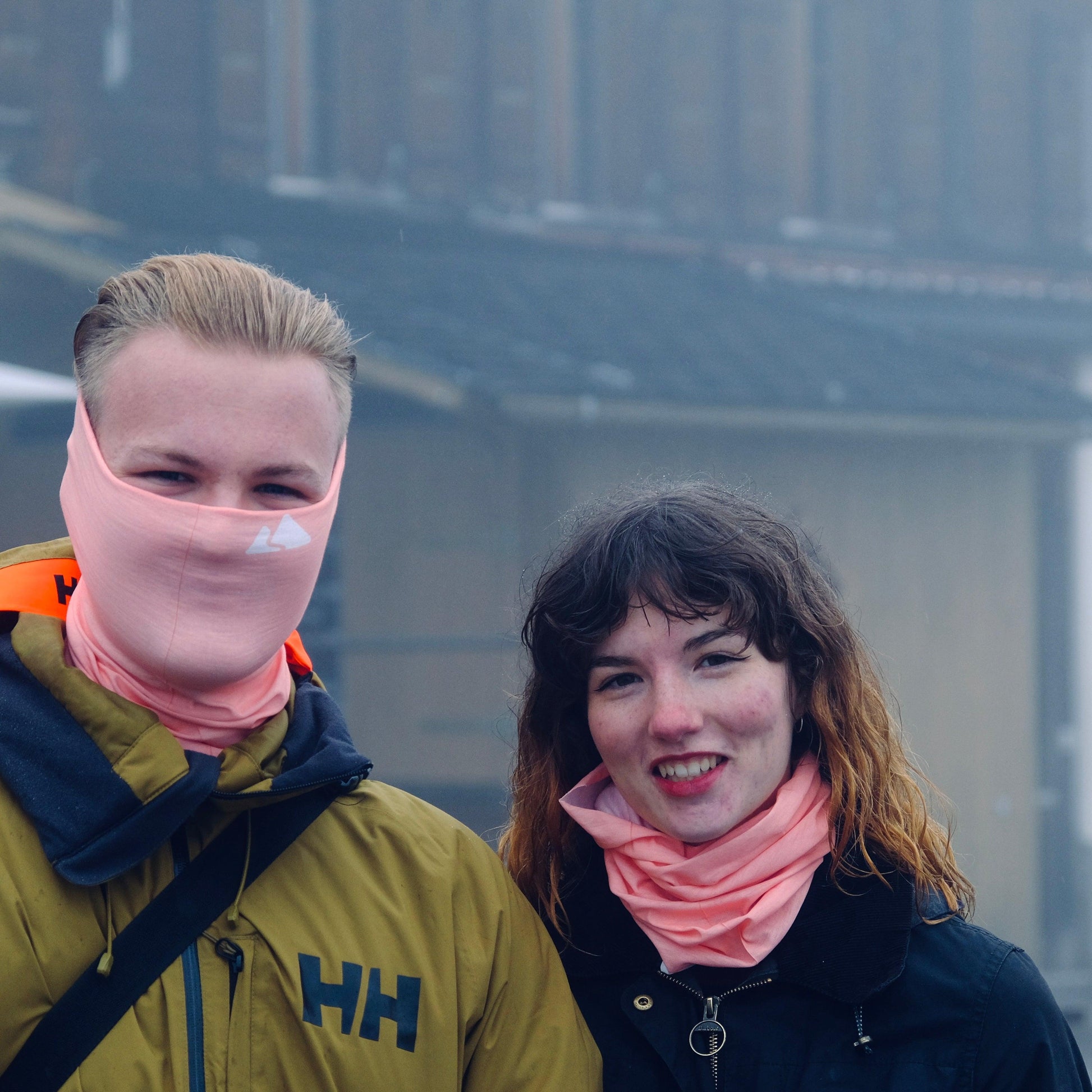 Neck buff gaiter displayed in a male and female model. With a white mountain logo. Blush Peach colour.  